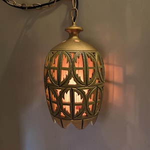 May include: A hanging pendant light fitting with a gold-toned, textured metal shade featuring a repeating circular cutout pattern. The lamp emits a warm, soft glow from within and is suspended by a gold-coloured metal chain and a black electrical cord.