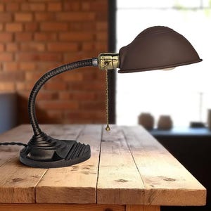 Puede incluir: Una lámpara de escritorio de estilo vintage con una pantalla marrón y un brazo de cuello de cisne negro flexible. La lámpara tiene una base negra y accesorios dorados. Está sobre una mesa de madera.