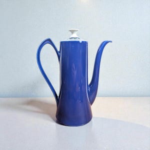 Puede incluir: Una cafetera de cerámica azul cobalto con un cuerpo alto y esbelto, un asa curvada y un pico largo y elegante. La tapa es blanca con un diseño decorativo verde y azul. Esta cafetera vintage es una adición elegante a cualquier cocina.