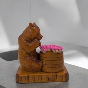 Peut inclure: Porte-allumettes en bois avec une figurine de ours sculptée tenant une allumette. Le porte-allumettes a un compartiment cylindrique rempli d'allumettes à tête rose.