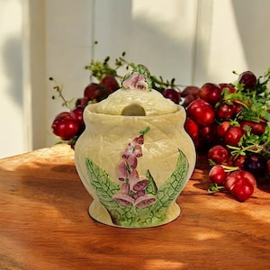 Peut inclure: Un sucrier en céramique crème avec un couvercle et une fente pour cuillère. Le bol est décoré d'un motif floral avec des fleurs violettes et des feuilles vertes. Le fond comprend des baies rouges et de la verdure.