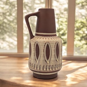 May include: A brown ceramic vase with a handle, featuring a cream-coloured, textured design with oval shapes. The vase is on a wooden surface, with a window and greenery in the background.