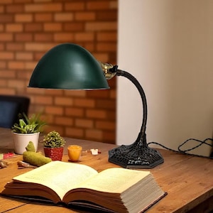Puede incluir: Una lámpara de escritorio verde con un brazo curvo y una base negra está sobre una mesa de madera. Un libro abierto está frente a la lámpara. El fondo incluye una pared de ladrillo y pequeñas plantas en macetas.