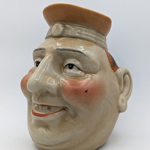 Könnte beinhalten: Eine Keramikkanne in Form eines Männerkopfes mit einem braunen Hut. Der Mann hat eine große Nase, rosige Wangen und ein breites Lächeln. Die Kanne ist in einem hellbraunen Ton mit einer craquelé-Oberfläche glasiert.