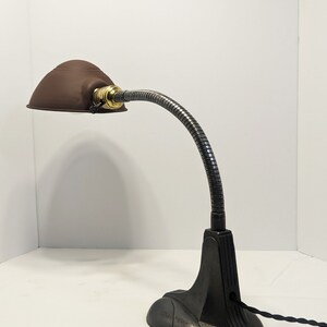 May include: A brown metal desk lamp with a flexible gooseneck arm and a black cast iron base. The lamp has a round shade and a brass switch.
