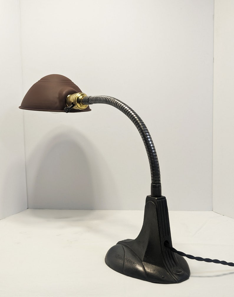 May include: A brown metal desk lamp with a flexible gooseneck arm and a black cast iron base. The lamp has a round shade and a brass switch.