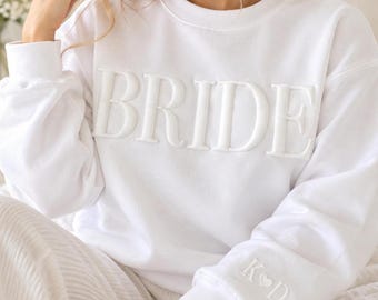 Puede incluir: Una sudadera blanca de cuello redondo con la palabra BRIDE en letras grandes, elevadas y tonales en el pecho. El puño de la manga izquierda muestra las iniciales bordadas K, un pequeño corazón y la letra D en hilo blanco.