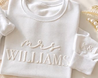 May include: A white crewneck sweatshirt featuring the embroidered text mrs. WILLIAMS in a raised white font on the chest and the initials K&I embroidered on the sleeve cuff, accompanied by two gold-toned pearl hair clips.
