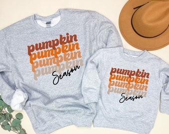 May include: Two grey sweatshirts with the text "pumpkin season" in orange, brown, and white lettering. The larger sweatshirt has four stacked "pumpkin" words, while the smaller sweatshirt has three.