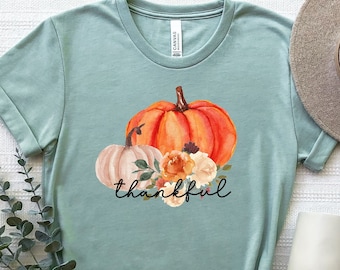 May include: A light blue t-shirt with a watercolor design of pumpkins and flowers. The text "thankful" is printed below the pumpkins.