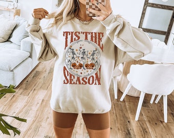 Könnte beinhalten: Ein beiger Pullover mit einem Grafikdruck von Skeletten, die Kürbisse halten, und dem Text "Tis the Season". Das Model trägt braune Radlerhosen und weiße Sneaker mit braun-schwarzen gestreiften Socken.
