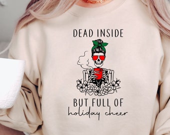May include: A beige sweatshirt with a graphic of a skeleton wearing sunglasses and a green headband, holding a red mug. The text "Dead Inside But Full of Holiday Cheer" is printed on the sweatshirt.