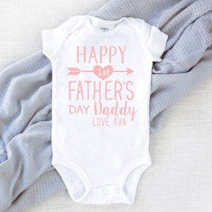 May include: White baby onesie with pink text that reads "Happy 1st Father's Day Daddy Love, Ava".