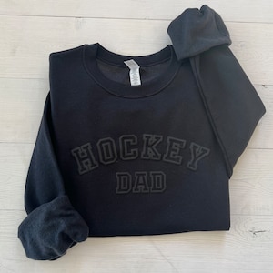 K&ouml;nnte beinhalten: Schwarzer Pullover mit dem Text "HOCKEY DAD" in wei&szlig;er Schrift mit schwarzem Umriss.