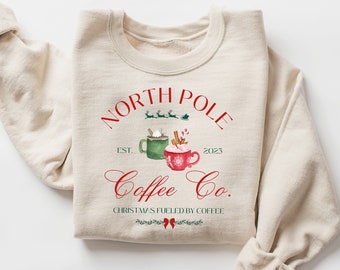 May include: A beige sweatshirt with a red and green graphic design that says "North Pole Coffee Co. Est. 2023. Christmas is fueled by coffee."