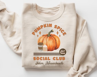 May include: A beige sweatshirt with a graphic design featuring a pumpkin, coffee cups, and the text "Pumpkin Spice Social Club Since 1992 Salem, Massachusetts".