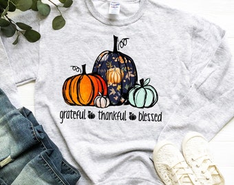 May include: A grey sweatshirt featuring a graphic of three pumpkins. The pumpkins are decorated with floral patterns and the text "grateful thankful blessed" is printed below them.