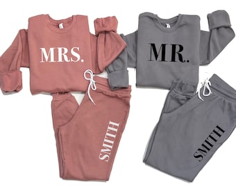 May include: Two matching sets of sweatpants and sweatshirts. The sweatshirts are grey and pink and have the words "MR." and "MRS." printed on them. The sweatpants are grey and pink and have the word "SMITH" printed on them.