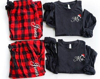May include: A set of matching pajamas with a red and black plaid pattern. The pajamas include a long-sleeved shirt and pants. The shirts have the text "Mr." and "Mrs." embroidered on them with a small red Santa hat graphic. The pants have the name "Jones" embroidered on them.