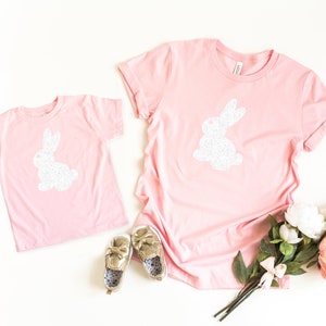 May include: Two pink t-shirts, one adult and one child size, each with a white glitter bunny design. Gold shoes and a bouquet of flowers are also in the image. The shirts are casual wear.
