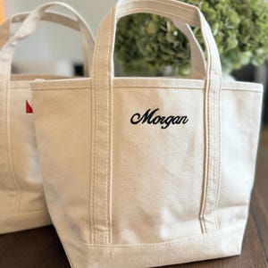 May include: An off-white canvas tote bag featuring two sturdy handles and the name Morgan embroidered in black script lettering on the front. The bag has a structured base and vertical stitched detailing along the front panel.