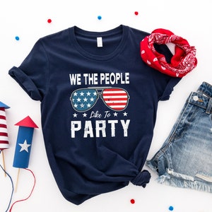 Może przedstawiać: Koszulka w kolorze granatowym z napisem "We the People Like to Party" i grafiką okular&oacute;w przeciwsłonecznych z wzorem amerykańskiej flagi. Koszulka leży na białym tle z czerwono-biało-niebieskimi konfetti.