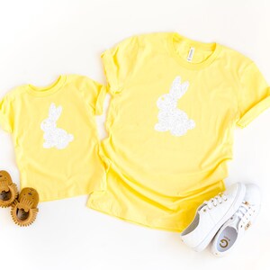 May include: Two yellow t-shirts, one adult and one child size, each featuring a white glitter bunny design. White sneakers and brown baby shoes are also in the image. The shirts are likely for Easter or spring.