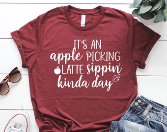 May include: A maroon t-shirt with the text "IT'S AN apple PICKING LATTE sippin' kinda day" in white. The shirt is paired with blue jeans, white sneakers, and sunglasses. A sprig of greenery is also visible.