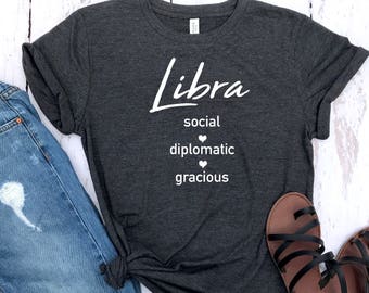 May include: A dark gray t-shirt with the word "Libra" in white lettering. The shirt also has the words "social", "diplomatic", and "gracious" in white lettering with a small heart symbol between each word.