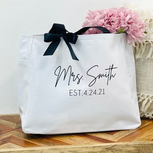 K&ouml;nnte beinhalten: Wei&szlig;e Canvas-Tragetasche mit schwarzen Henkeln und einer schwarzen Schleife. Die Tasche tr&auml;gt den Aufdruck "Mrs. Smith EST. 4.24.21".