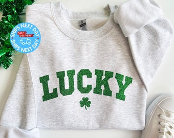 Op de afbeelding: Een grijs sweatshirt met het woord "LUCKY" in groene glitterletters en een groen klavertje vier onderaan.