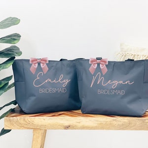May include: Two gray tote bags with pink bows and the names "Emily Bridesmaid" and "Megan Bridesmaid" printed on them.