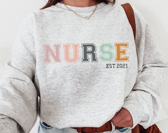 May include: A gray sweatshirt with the word "NURSE" in colorful letters and the text "EST. 2021" below it.