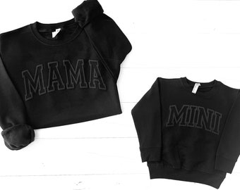 May include: Two black sweatshirts with the words "MAMA" and "MINI" in a bold, outlined font. The sweatshirts are laid out on a white wooden surface.