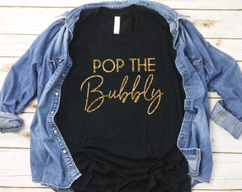 May include: Black t-shirt with gold glitter lettering that reads "Pop the Bubbly". The shirt is folded and laid on a white wooden surface.