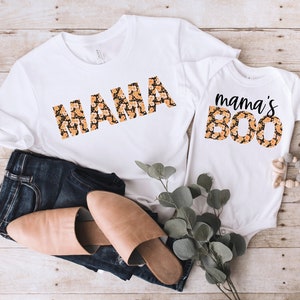 May include: A white t-shirt with the word "MAMA" in black letters with a floral pattern. A white onesie with the text "mama's BOO" in black letters with a floral pattern.