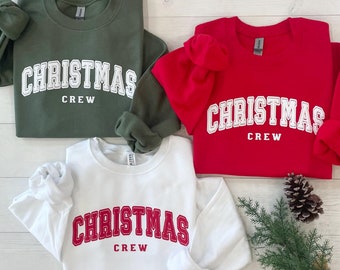 May include: Three crewneck sweatshirts in olive green, bright red, and white. Each sweatshirt features the text "CHRISTMAS CREW" printed in a collegiate-style font. The olive green and red shirts have white text, while the white shirt has red text.