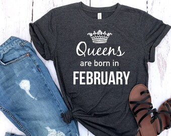 May include: A dark gray t-shirt with a white crown and the text "Queens are born in February". The shirt is laying on a wooden surface with a pair of blue jeans and sandals.