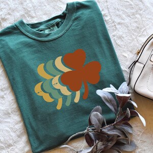 Może przedstawiać: Zielony t-shirt z nadrukiem retro czterolistnej koniczyny w odcieniach zieleni, ż&oacute;łci i pomarańczy. Koniczyna jest umieszczona centralnie na koszulce.
