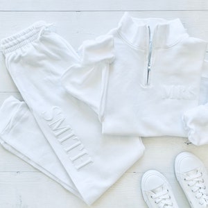 Op de afbeelding: Witte sweatpants met het woord "SMITH" geborduurd aan de zijkant, een wit kwart-rits sweatshirt met het woord "MRS" geborduurd aan de voorkant en twee paar witte sneakers.
