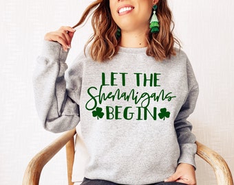 Könnte beinhalten: Grauer Pullover mit dem Text "Let the Shenanigans Begin" in grüner Schrift mit Kleeblatt-Symbolen auf beiden Seiten des Textes.