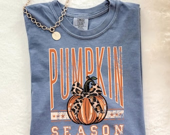 May include: A blue Comfort Colors t-shirt with the words "PUMPKIN SEASON" in orange and a pumpkin graphic. A gold chain necklace with a round pendant is draped over the shirt.