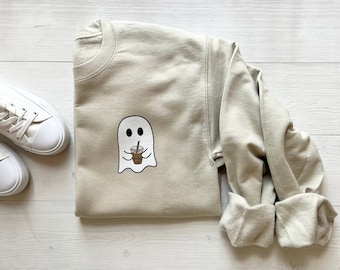 May include: A light beige sweatshirt with a ghost holding a cup of coffee printed on the front.