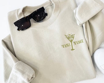 Könnte beinhalten: Ein hellbeiger Pullover mit einem grün gestickten Martini-Glas und dem Text "Tini Time".