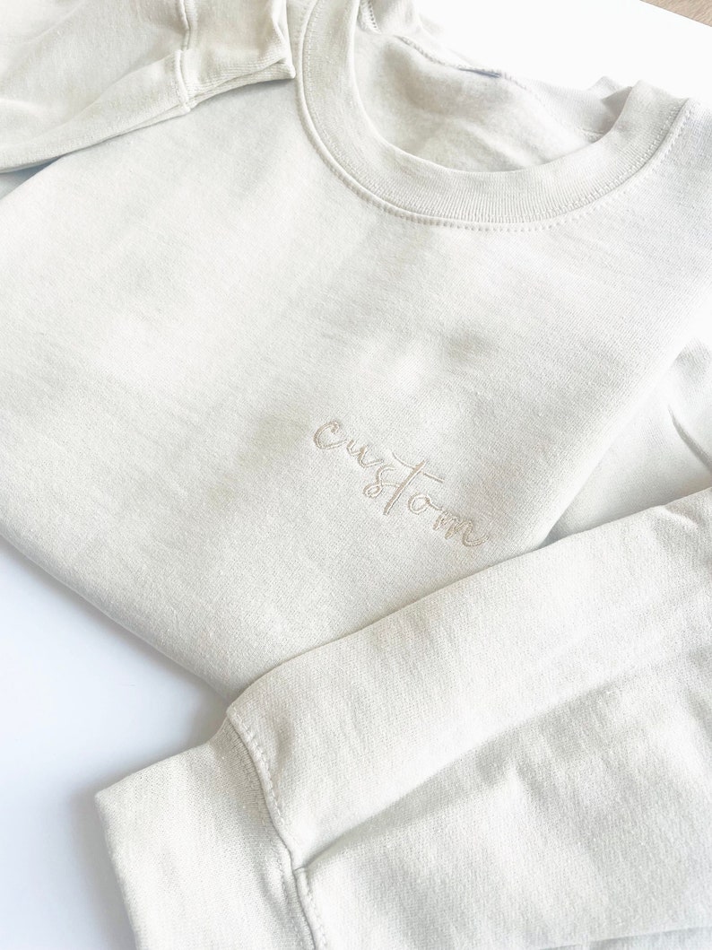 K&ouml;nnte beinhalten: Ein hellbeiger Pullover mit dem Wort "custom" in kursiv auf der Vorderseite gestickt.