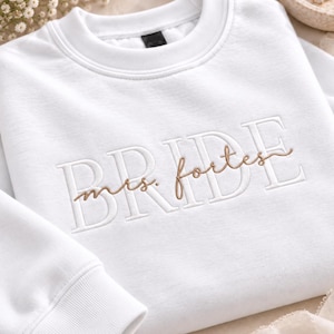 May include: A white crewneck sweatshirt featuring the word BRIDE embroidered in large white block letters across the chest, with the text mrs. fortes embroidered in a smaller, cursive brown script layered over the center of the word.