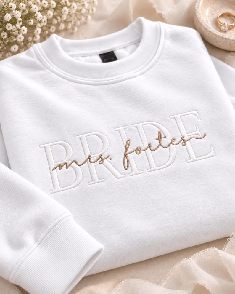 May include: A white crewneck sweatshirt featuring the word BRIDE embroidered in large white block letters across the chest, with the text mrs. fortes embroidered in a smaller, cursive brown script layered over the center of the word.