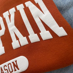 Op de afbeelding: Een roestoranje sweatshirt met grote witte blokletters die het woord PUMPKIN vormen. Onder de hoofdtekst bevindt zich een witte, afgeronde rechthoekige patch met het woord SEASON in roestoranje letters. De stof heeft een zichtbare textuur.