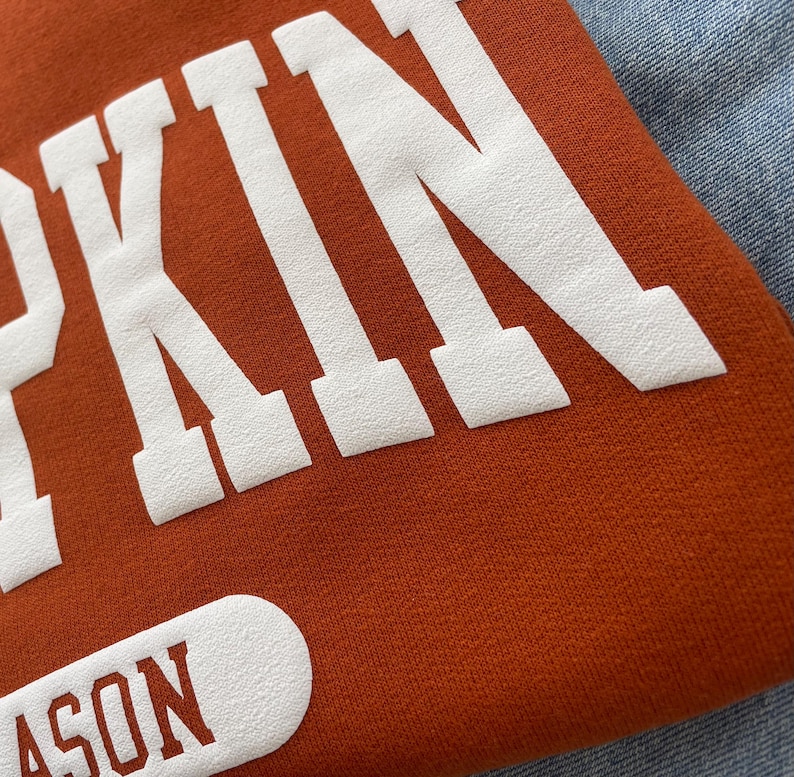 Op de afbeelding: Een roestoranje sweatshirt met grote witte blokletters die het woord PUMPKIN vormen. Onder de hoofdtekst bevindt zich een witte, afgeronde rechthoekige patch met het woord SEASON in roestoranje letters. De stof heeft een zichtbare textuur.