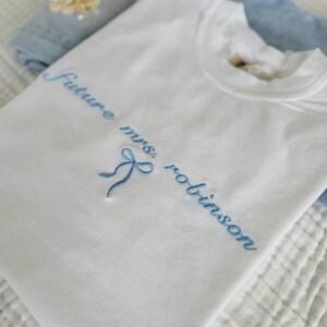 Op de afbeelding: Wit T-shirt met blauwe borduurwerk dat "Future Mrs. Robinson" zegt met een blauwe strik.